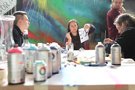 Junge Menschen sitzen an einem Tisch und malen, auf dem Tisch stehen Spraydosen und Stifte