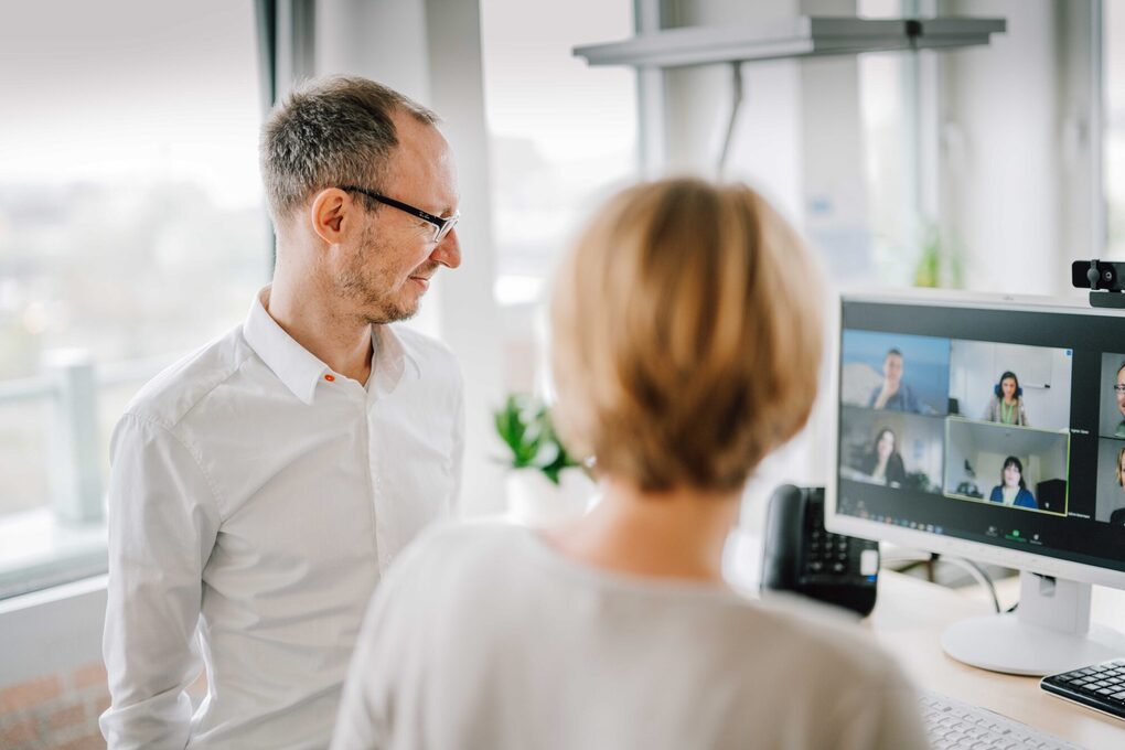 Zwei Personen stehen vor einem Bildschirm auf dem eine Videokonferenz zu sehen ist