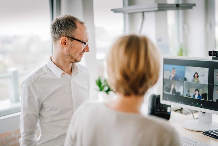 Zwei Personen stehen vor einem Bildschirm auf dem eine Videokonferenz zu sehen ist