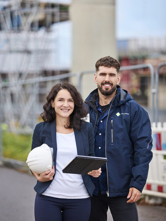 Zwei Personen stehen auf einer Baustelle, die Frau hat eine Mappe und einen Schutzhelm in den Händen