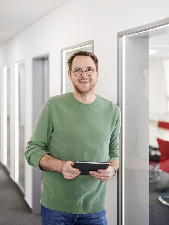 Ein Mann hat ein Tablet in der Hand und lehnt im Flur eines Bürogebäudes
