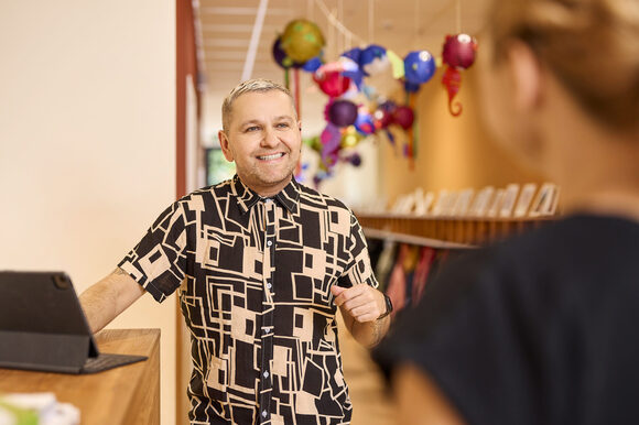 Ein junger Mann steht vor einem Tablet im Flur einer Kita und redet mit einer anderen Person