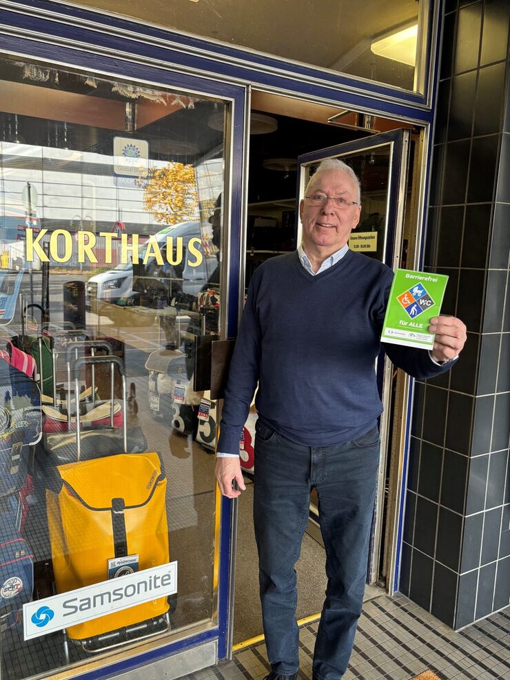 Eine Person steht vor dem Eingang von Lederwaren Korthaus und hält das Barrierefrei Siegel in der Hand