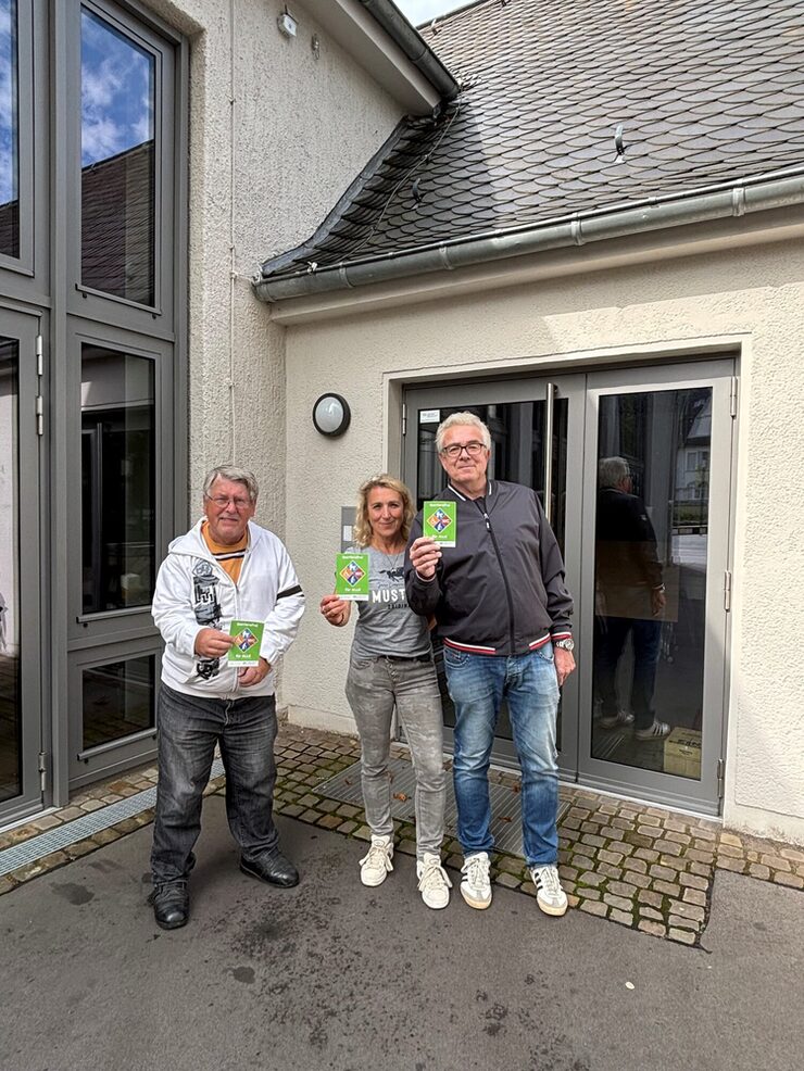 Drei Personen stehen vor der Mehrzweckhalle Opladen und halten das Barrierefrei-Siegel in der Hand